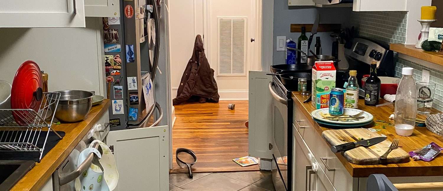A view through a galley kitchen with food and dishtowels on the floor, prep mess on the counter, a cupboard door left open, and bibs hanging from the oven door handle.